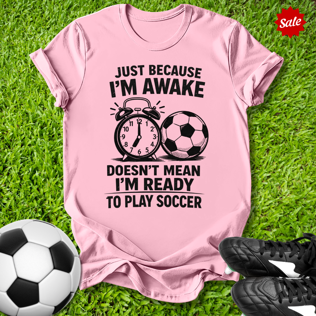 Pink t-shirt with soccer-themed text on grass with soccer balls and shoes.