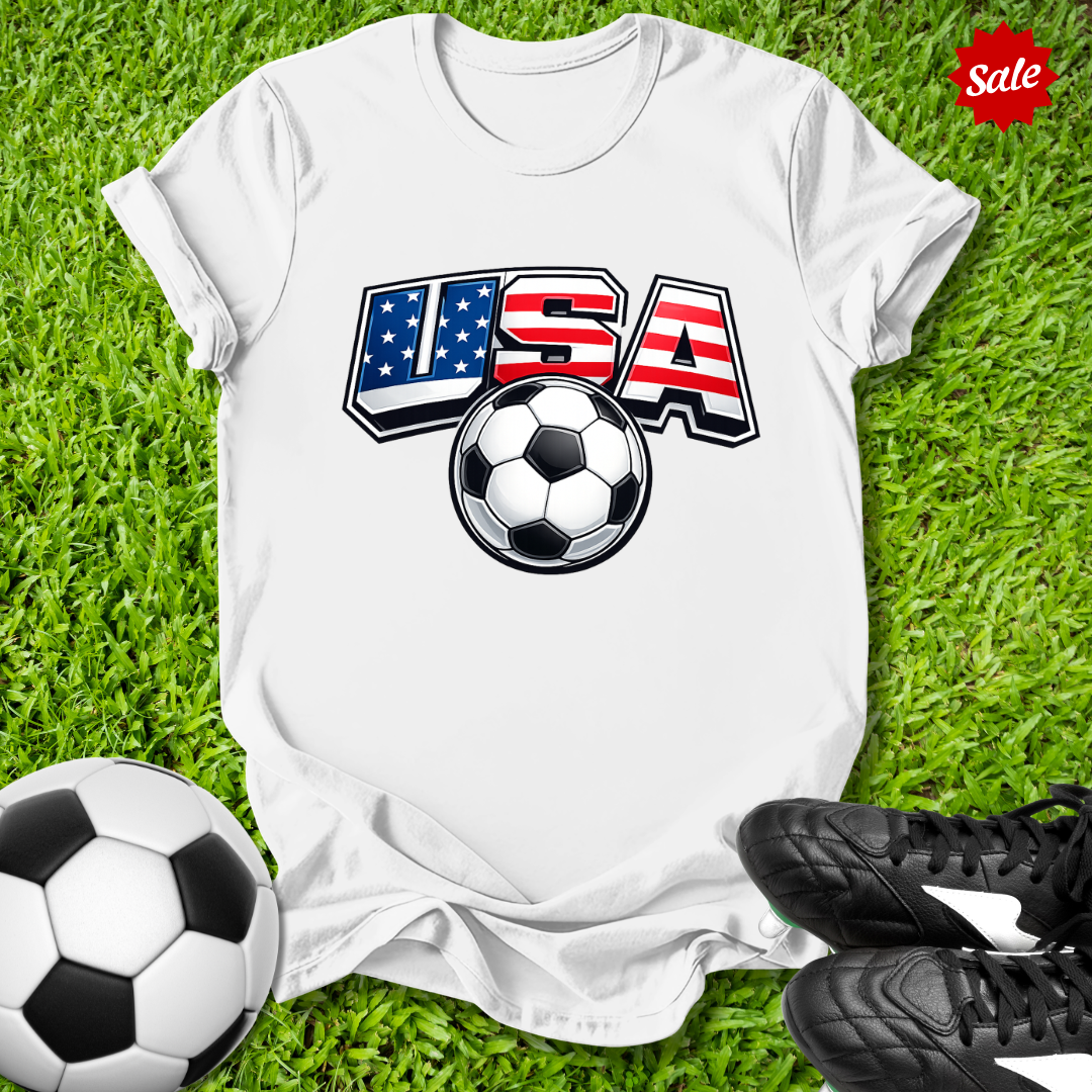 White t-shirt with USA and soccer ball design on grass with soccer balls and shoes.