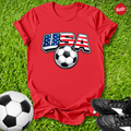 Red t-shirt with USA and soccer ball design on grass with soccer ball and shoes.