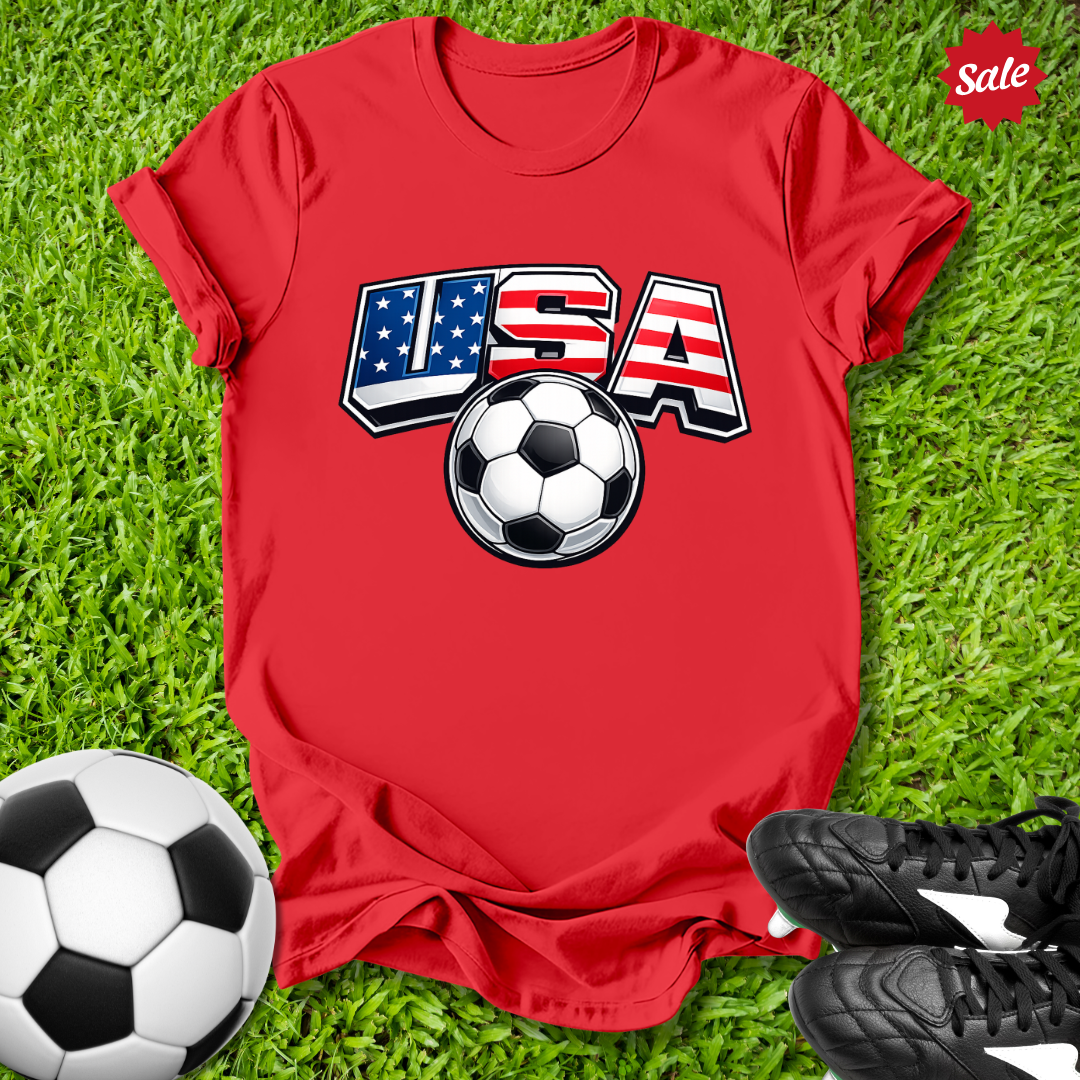 Red t-shirt with USA and soccer ball design on grass with soccer ball and shoes.