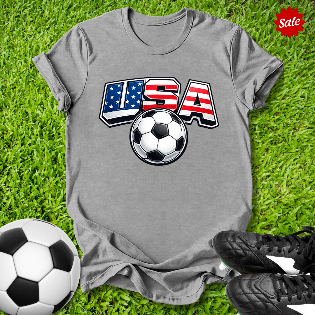 Gray t-shirt with USA soccer design on grass with soccer balls and shoes.