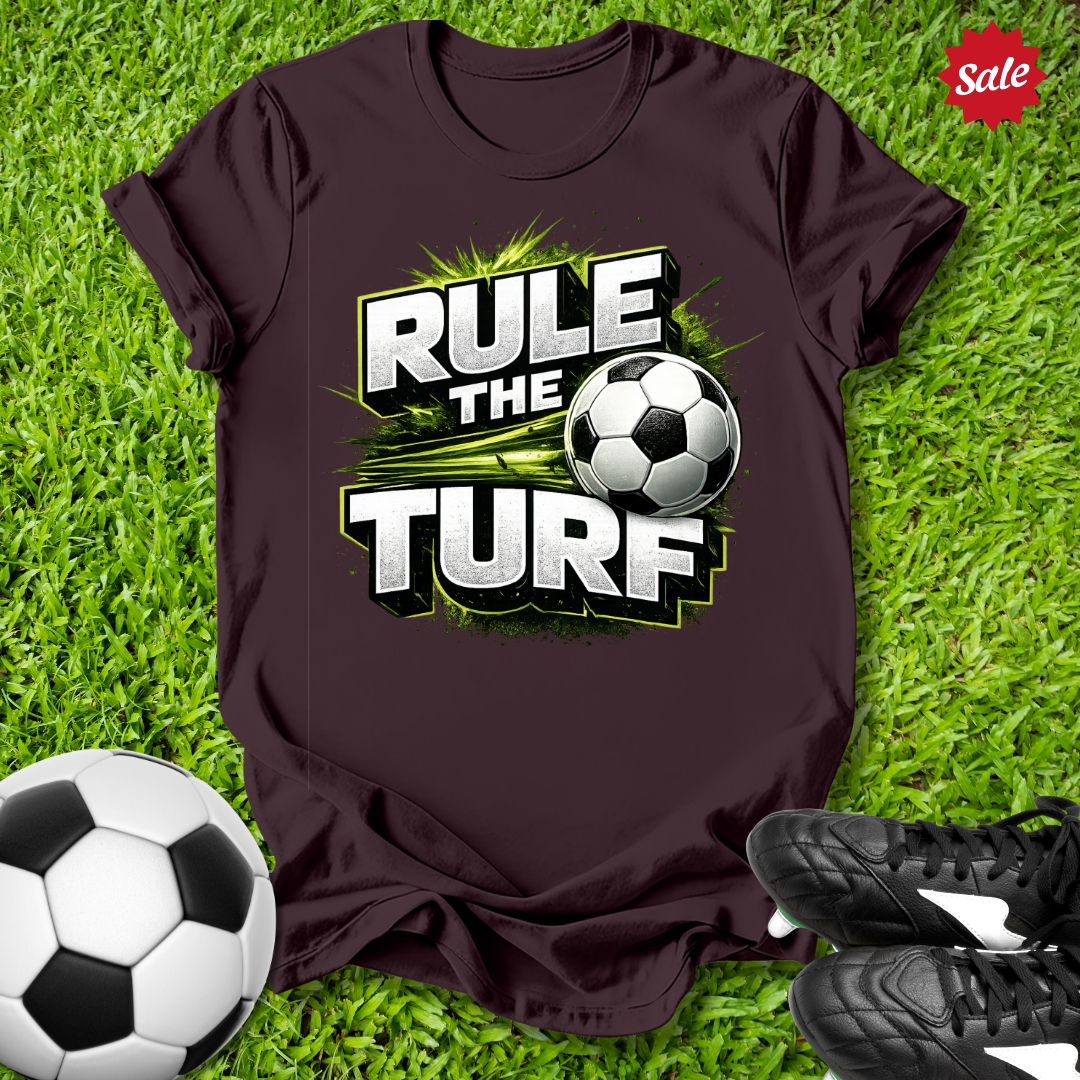 Brown t-shirt with 'Rule the Turf' graphic on grass with soccer balls and shoes.