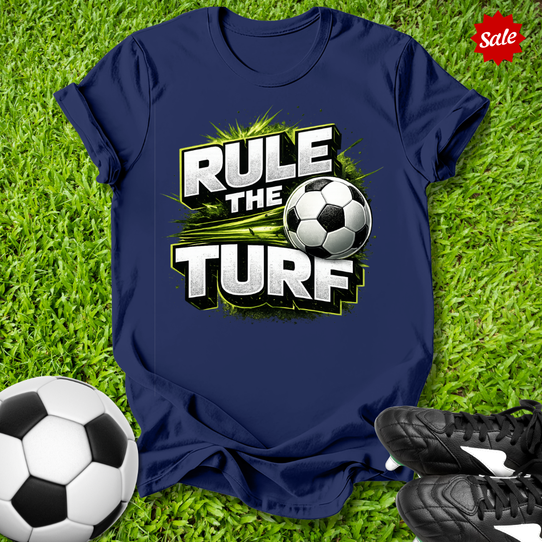Navy t-shirt with 'Rule the Turf' design on grass with soccer ball and shoes.