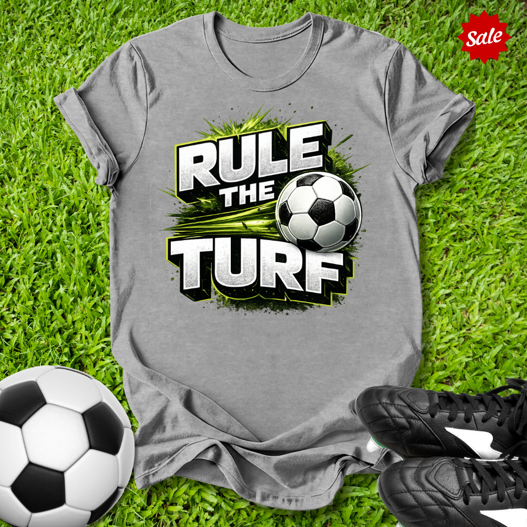Gray t-shirt with 'Rule the Turf' design on grass with soccer balls and shoes.