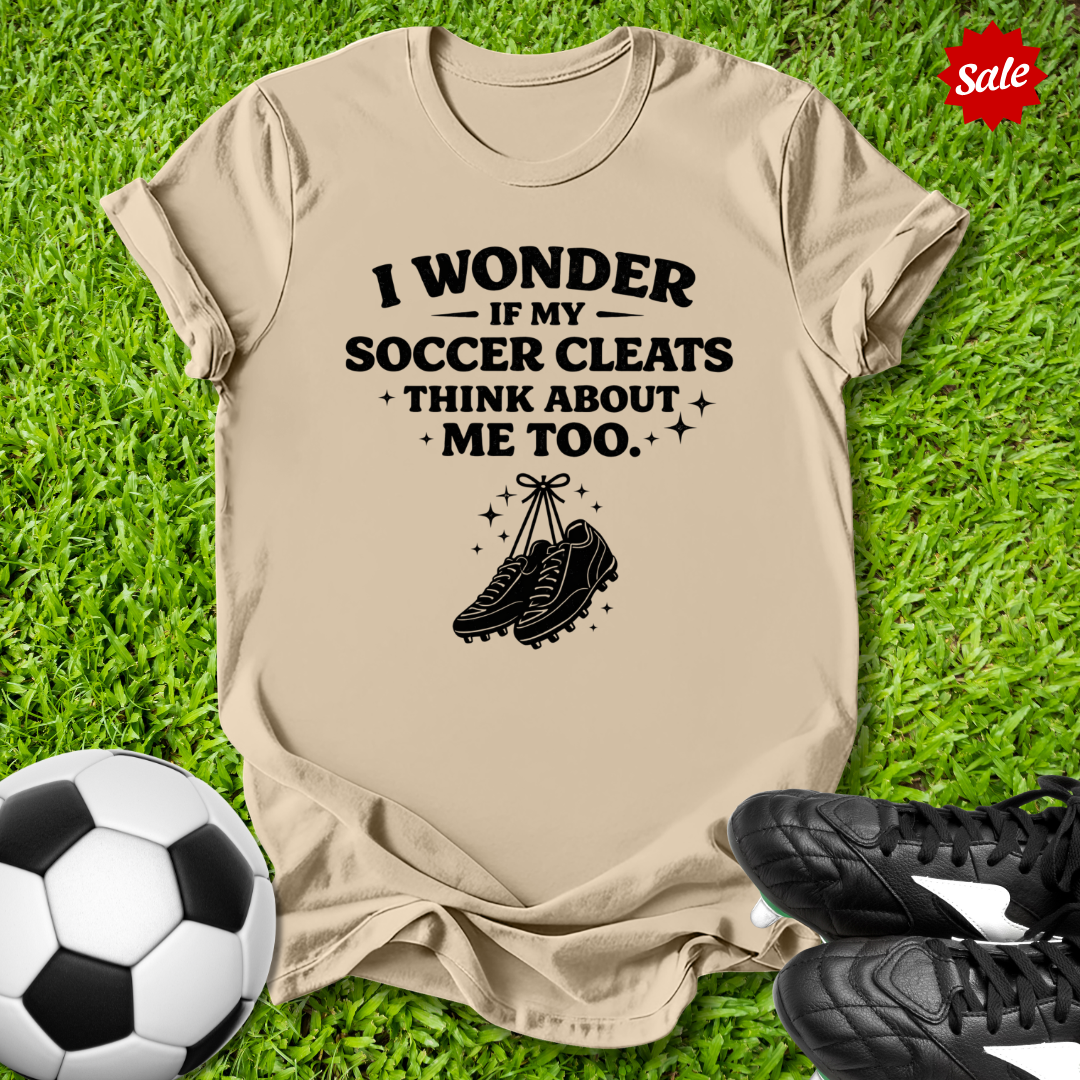 Beige t-shirt with soccer-themed text and graphics on grass with soccer ball and cleats.