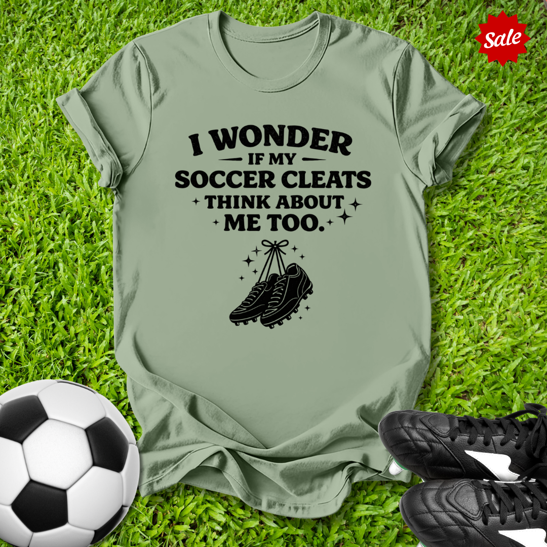 Green t-shirt with soccer-themed text and graphics on grass with soccer ball and cleats.