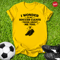 Yellow t-shirt with soccer-themed text and graphics on grass with soccer ball and cleats.