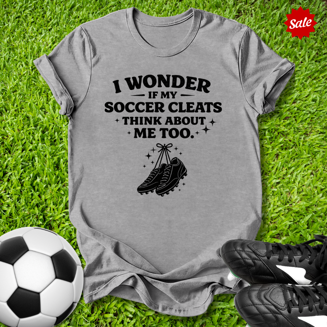 Gray t-shirt with soccer-themed text and graphics on grass with soccer ball and cleats.