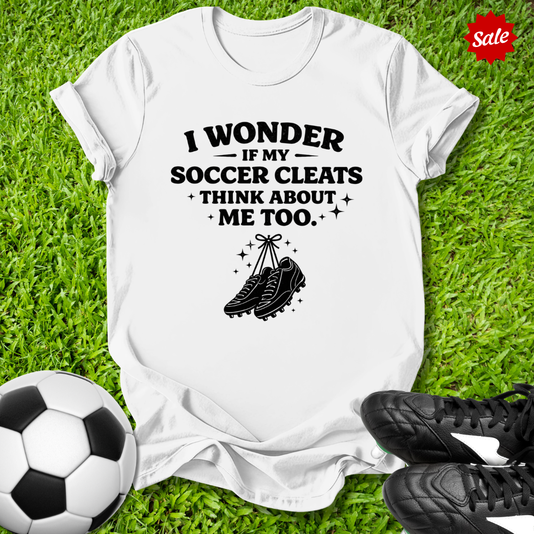 White t-shirt with soccer-themed text and graphics on grass with soccer ball and cleats.