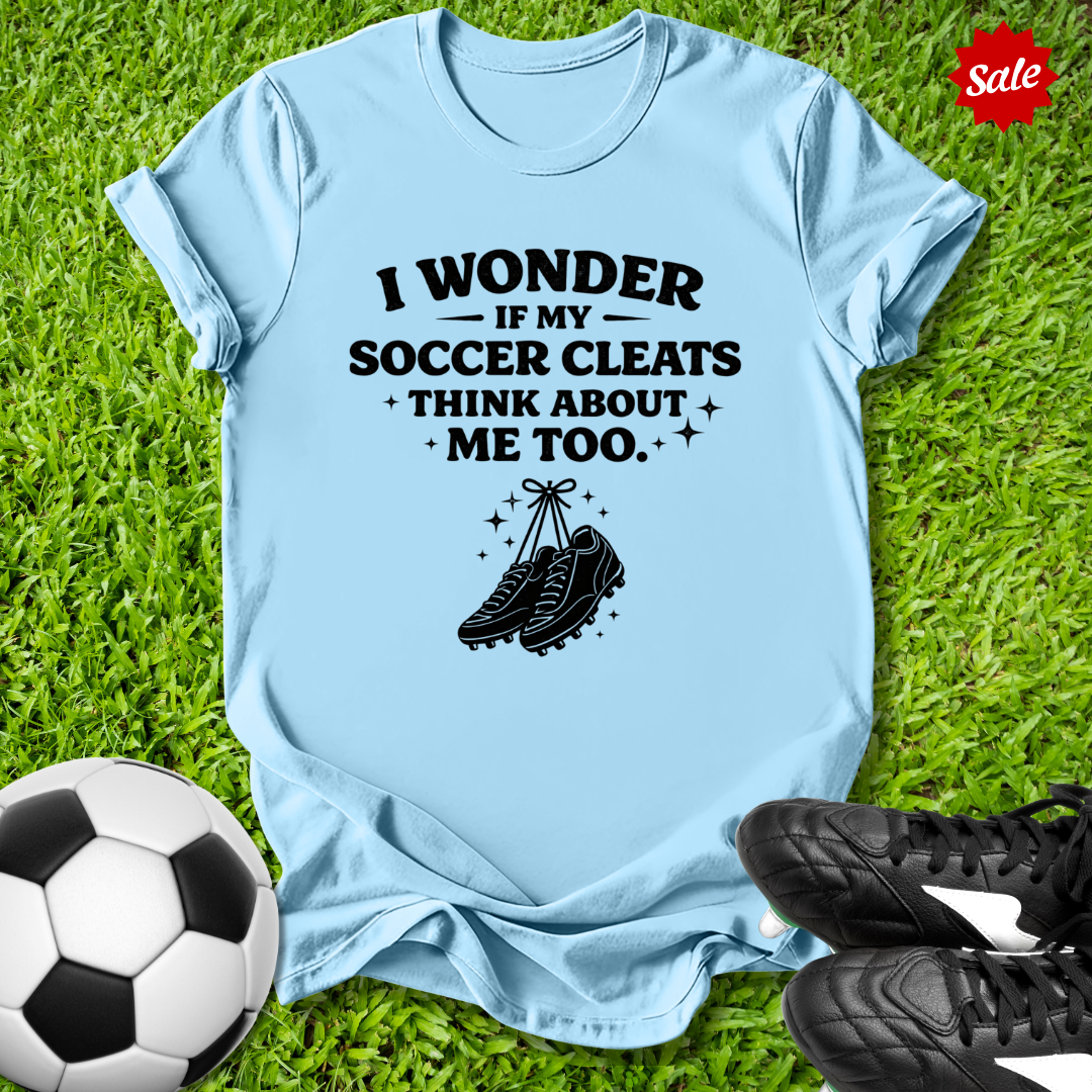 Light blue t-shirt with soccer-themed text and graphics on grass with soccer ball and cleats.