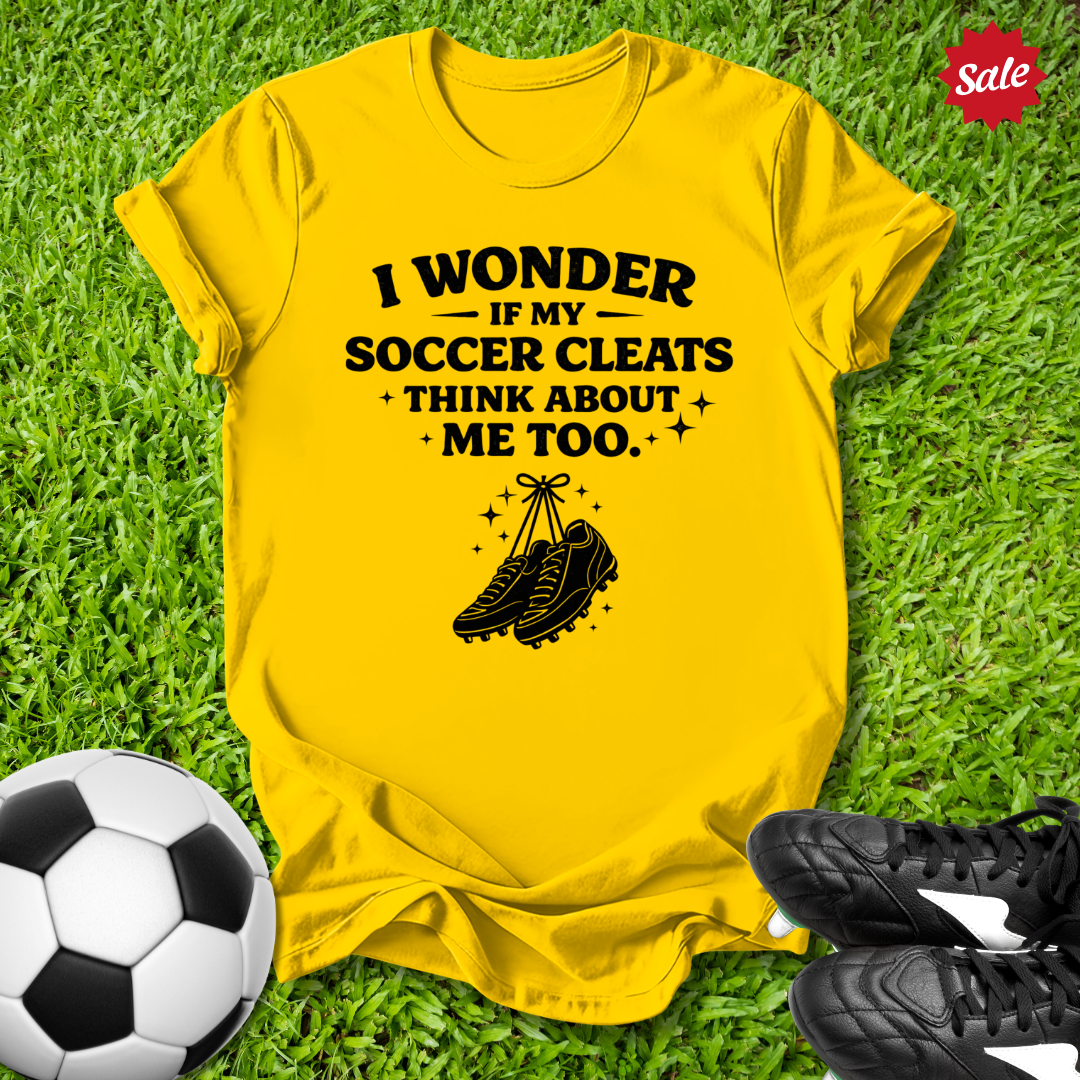 Yellow t-shirt with soccer-themed text and graphics on grass with soccer ball and cleats.