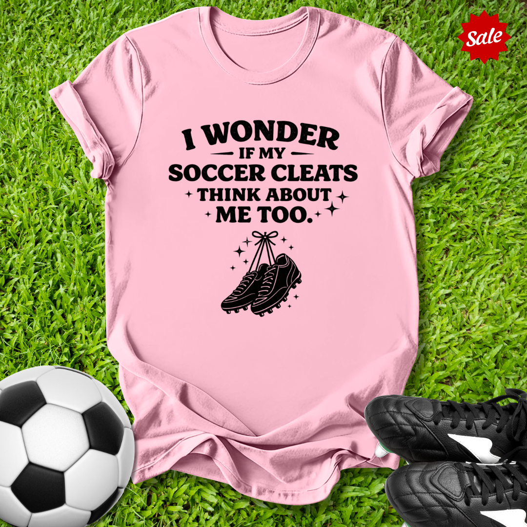 Pink t-shirt with soccer-themed text and graphics on grass with soccer ball and cleats.
