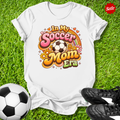 White t-shirt with 'In My Soccer Mom Era' design on grass with soccer ball and shoes.