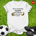 White t-shirt with humorous my car smells like cleats and a banana from last season text on grass with soccer ball and cleats