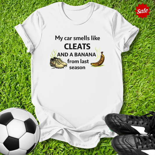 White t-shirt with humorous my car smells like cleats and a banana from last season text on grass with soccer ball and cleats