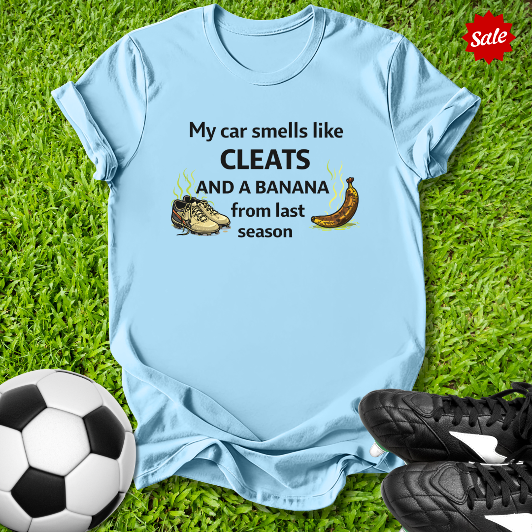 Light blue t-shirt with humorous  my car smells like cleats and a banana from last season text on grass with soccer ball and cleats