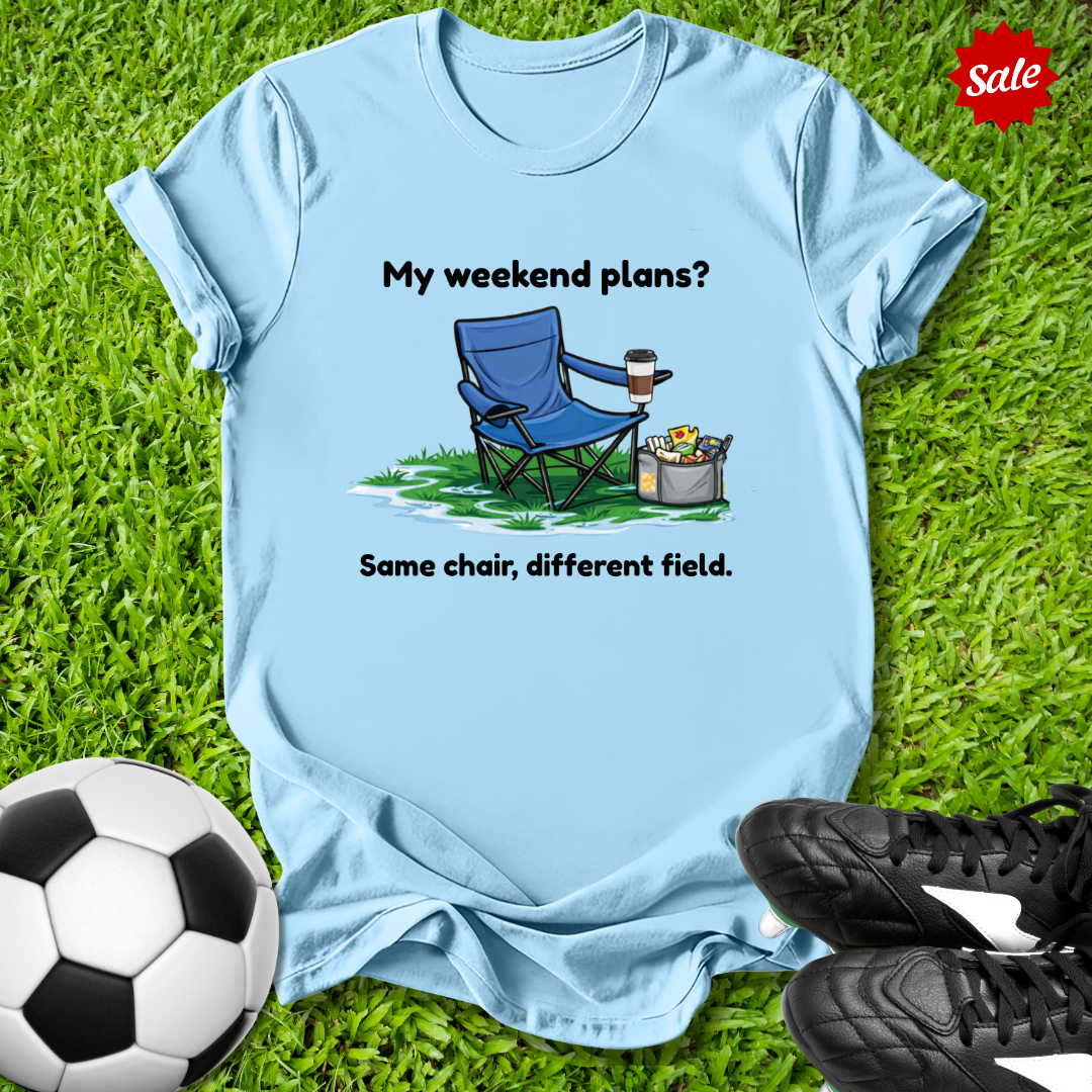 Light blue t-shirt with a graphic and text on grass with soccer ball and shoes