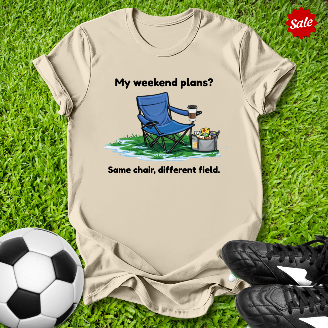 Beige t-shirt with a graphic of a blue chair and text on grass with soccer ball and shoes.