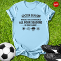 Light blue t-shirt with soccer-themed text on grass with soccer ball and shoes