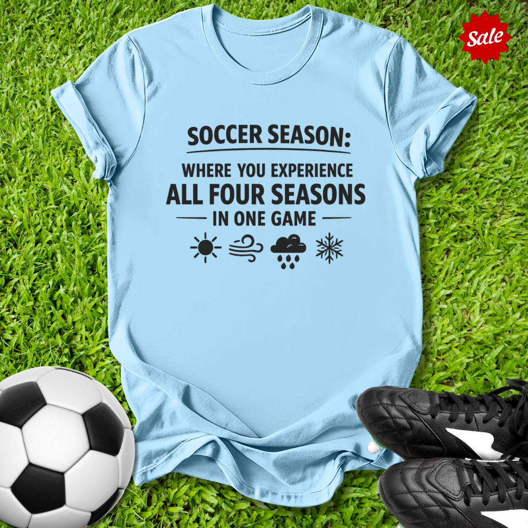Light blue t-shirt with soccer-themed text on grass with soccer ball and shoes