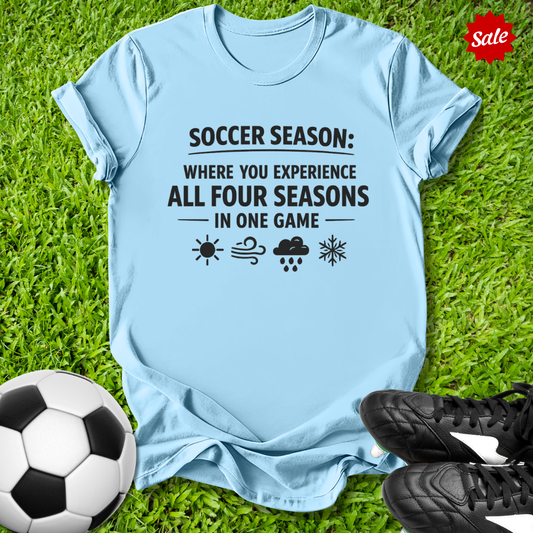 Light blue t-shirt with soccer-themed text on grass with soccer ball and shoes