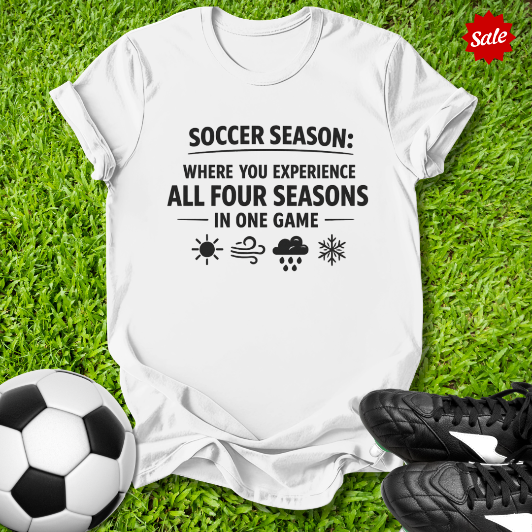 White t-shirt with soccer-themed text on grass with soccer ball and shoes