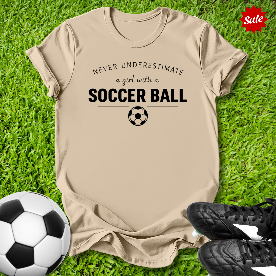 Beige t-shirt with 'Never underestimate a girl with a soccer ball' text on grass with soccer ball and shoes.