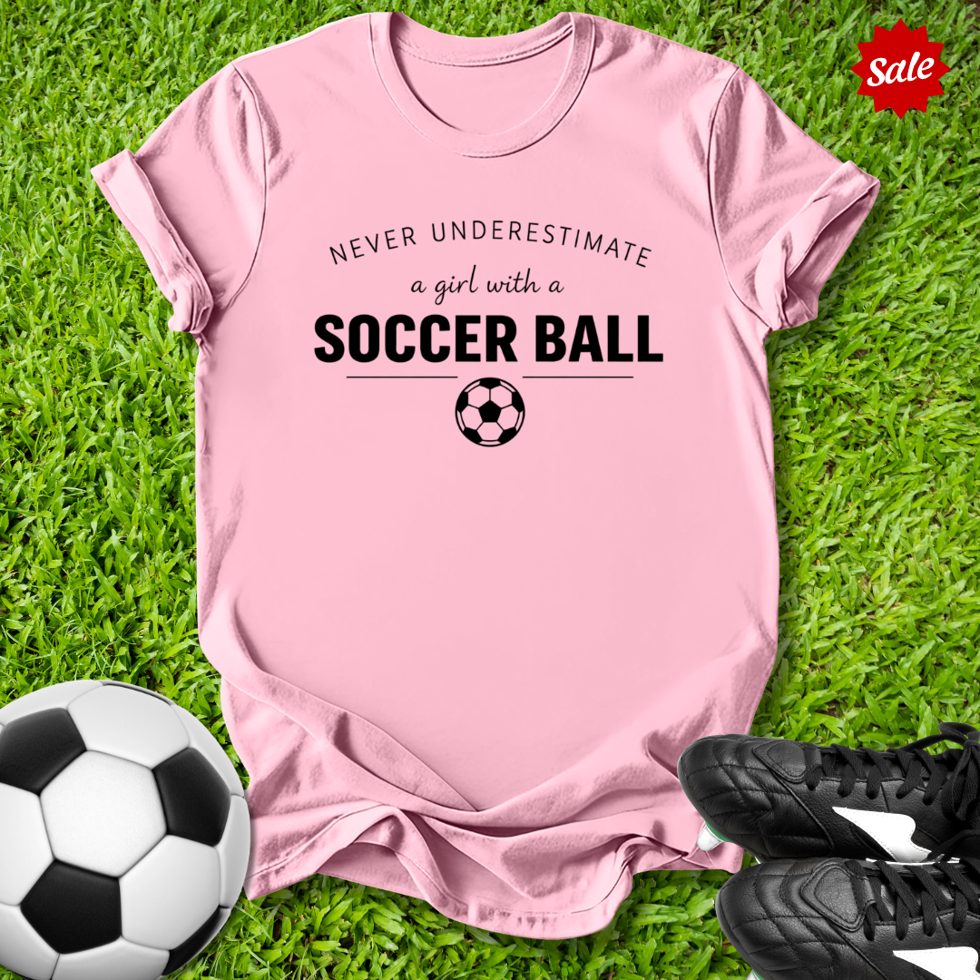 Pink t-shirt with 'Never Underestimate a Girl with a Soccer Ball' text on grass with soccer ball and shoes.