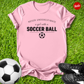Pink t-shirt with 'Never Underestimate a Girl with a Soccer Ball' text on grass with soccer ball and shoes.