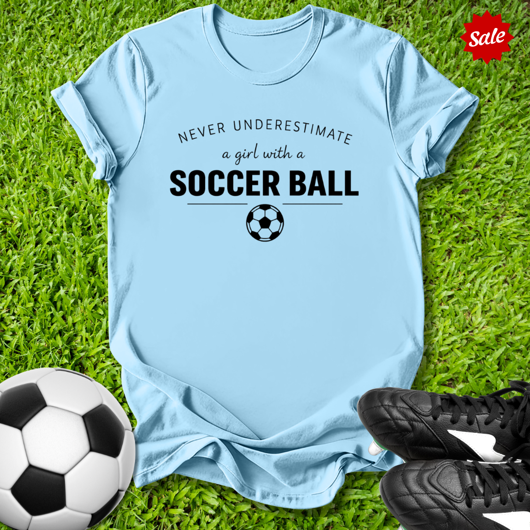 Light blue t-shirt with 'Never underestimate a girl with a soccer ball' text on grass with soccer ball and shoes.