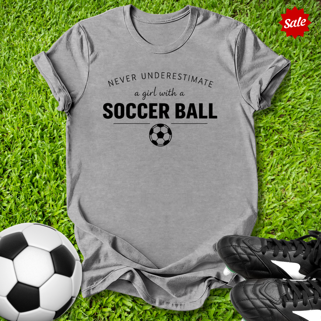 Gray t-shirt with 'Never underestimate a girl with a soccer ball' text on grass with soccer ball and shoes.