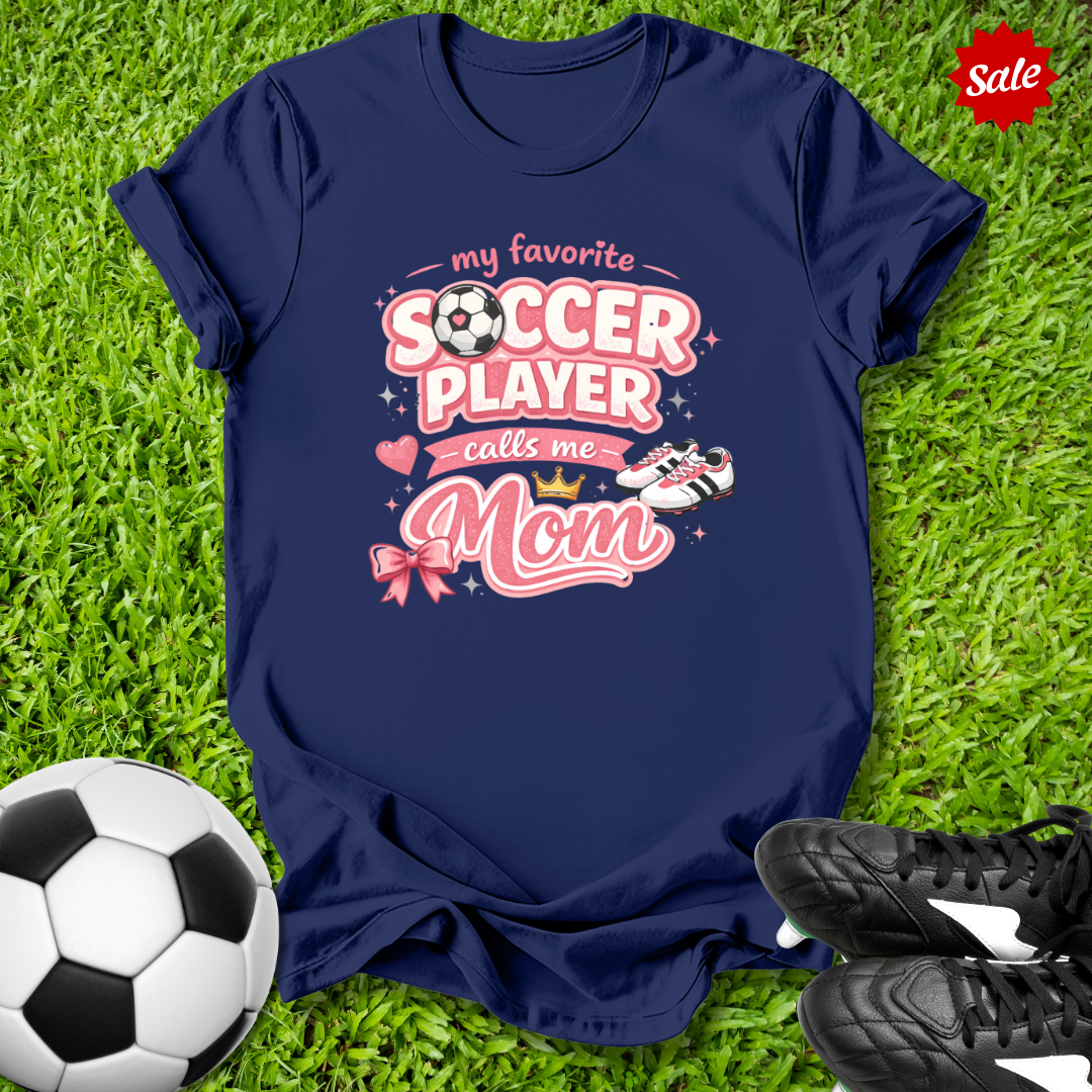 Navy blue t-shirt with pink soccer-themed design and My Favorite Player Calls Me Mom text  on grass with soccer ball and shoes