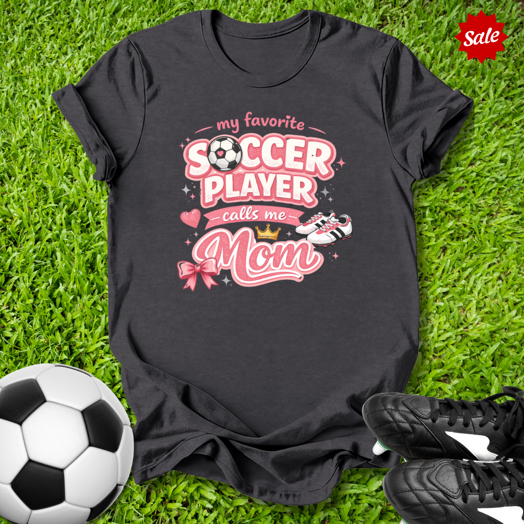 Black t-shirt with pink soccer-themed design and My Favorite Player Calls Me Mom text on grass with soccer ball and shoes