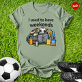 Green t-shirt with graphic and text 'I used to have weekends' on grass with soccer ball and shoes.