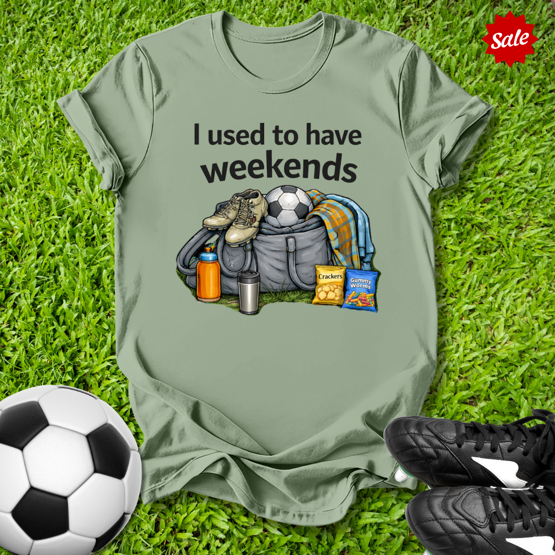 Green t-shirt with graphic and text 'I used to have weekends' on grass with soccer ball and shoes.