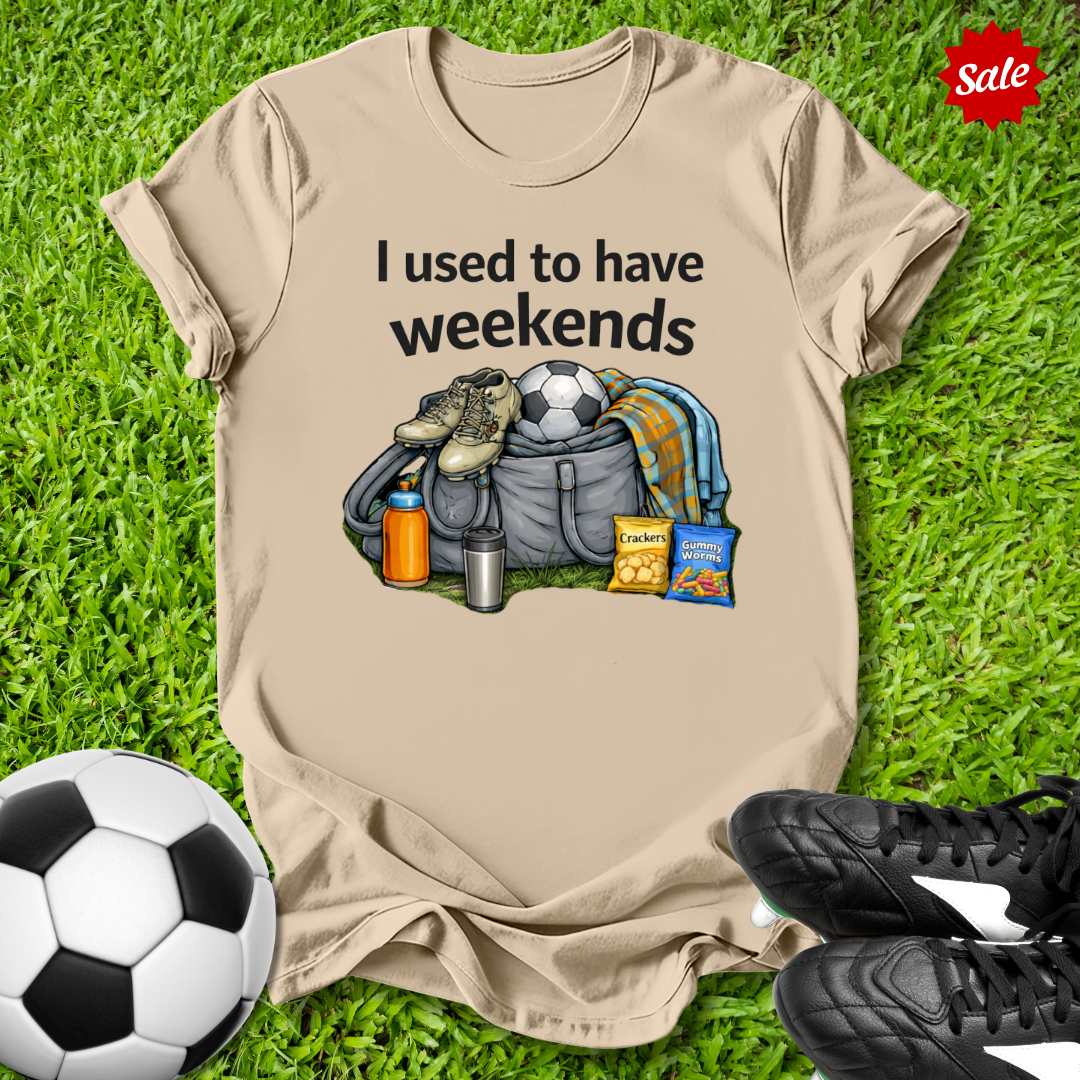 Beige t-shirt with graphic and text on grass with soccer ball and shoes
