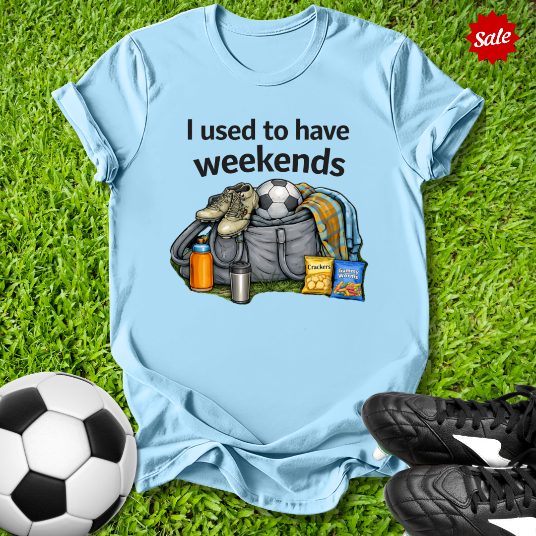 Light blue t-shirt with graphic and text 'I used to have weekends' on grass with soccer ball and shoes.