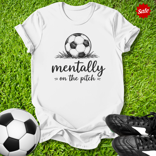 White t-shirt with soccer ball graphic and text 'mentally on the pitch' on grass with soccer balls and shoes.