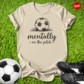 Beige t-shirt with soccer ball graphic and text on grass with soccer balls and shoes.