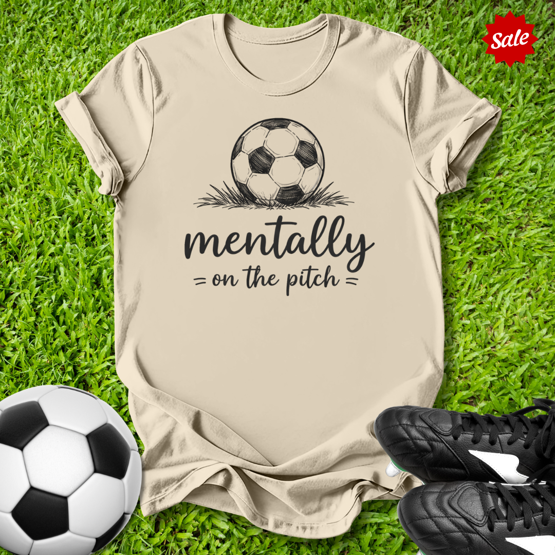 Beige t-shirt with soccer ball graphic and text on grass with soccer balls and shoes.