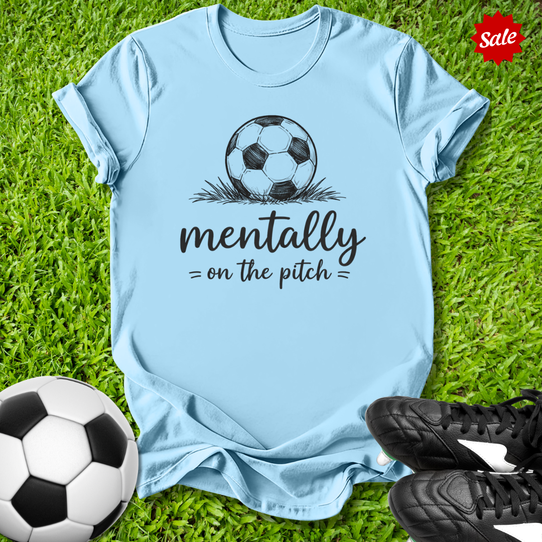 Light blue t-shirt with soccer ball graphic and text on grass with soccer ball and shoes.