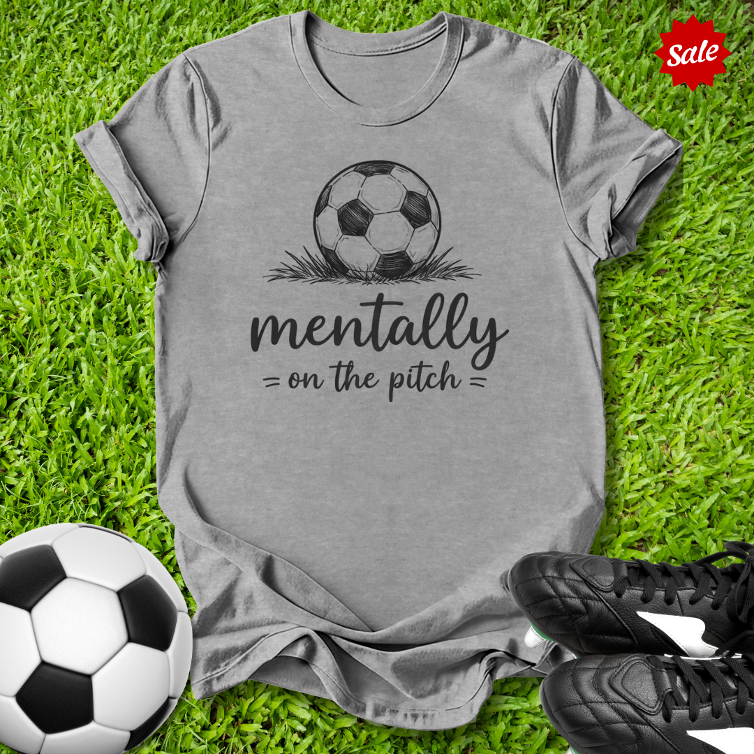 Gray t-shirt with soccer ball graphic and text on grass with soccer balls and shoes.