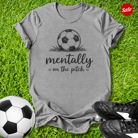 Gray t-shirt with soccer ball graphic and text on grass with soccer balls and shoes.