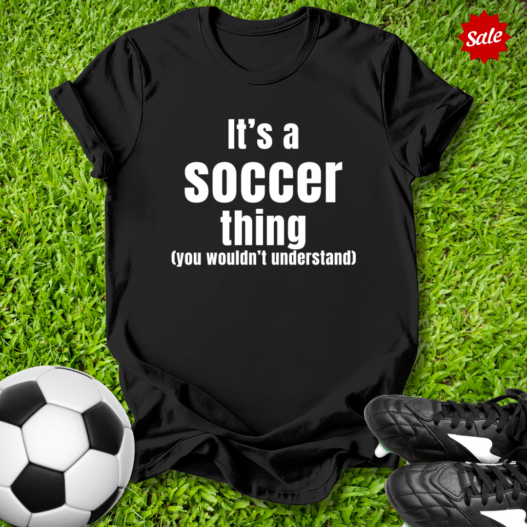 Black t-shirt with 'It's a soccer thing (you wouldn't understand)' text on grass with soccer ball and shoes.