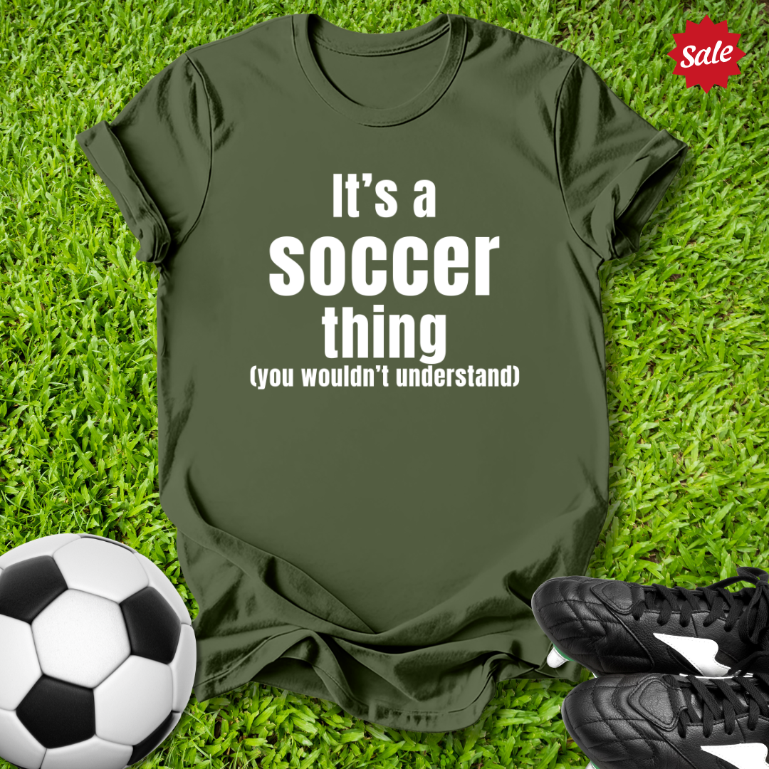Green t-shirt with 'It's a soccer thing (you wouldn't understand)' text on grass with soccer ball and shoes.