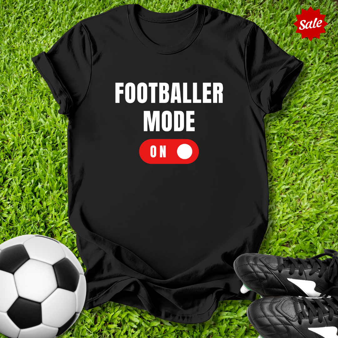 Black t-shirt with 'FOOTBALLER MODE ON' text on grass with soccer ball and shoes