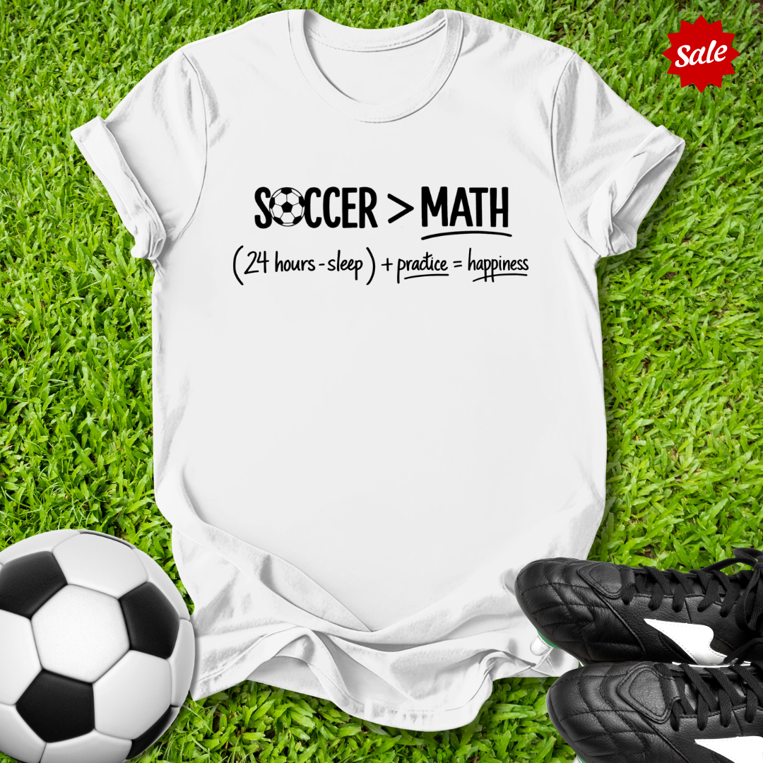 White t-shirt with 'Soccer > Math' text on grass with soccer ball and shoes