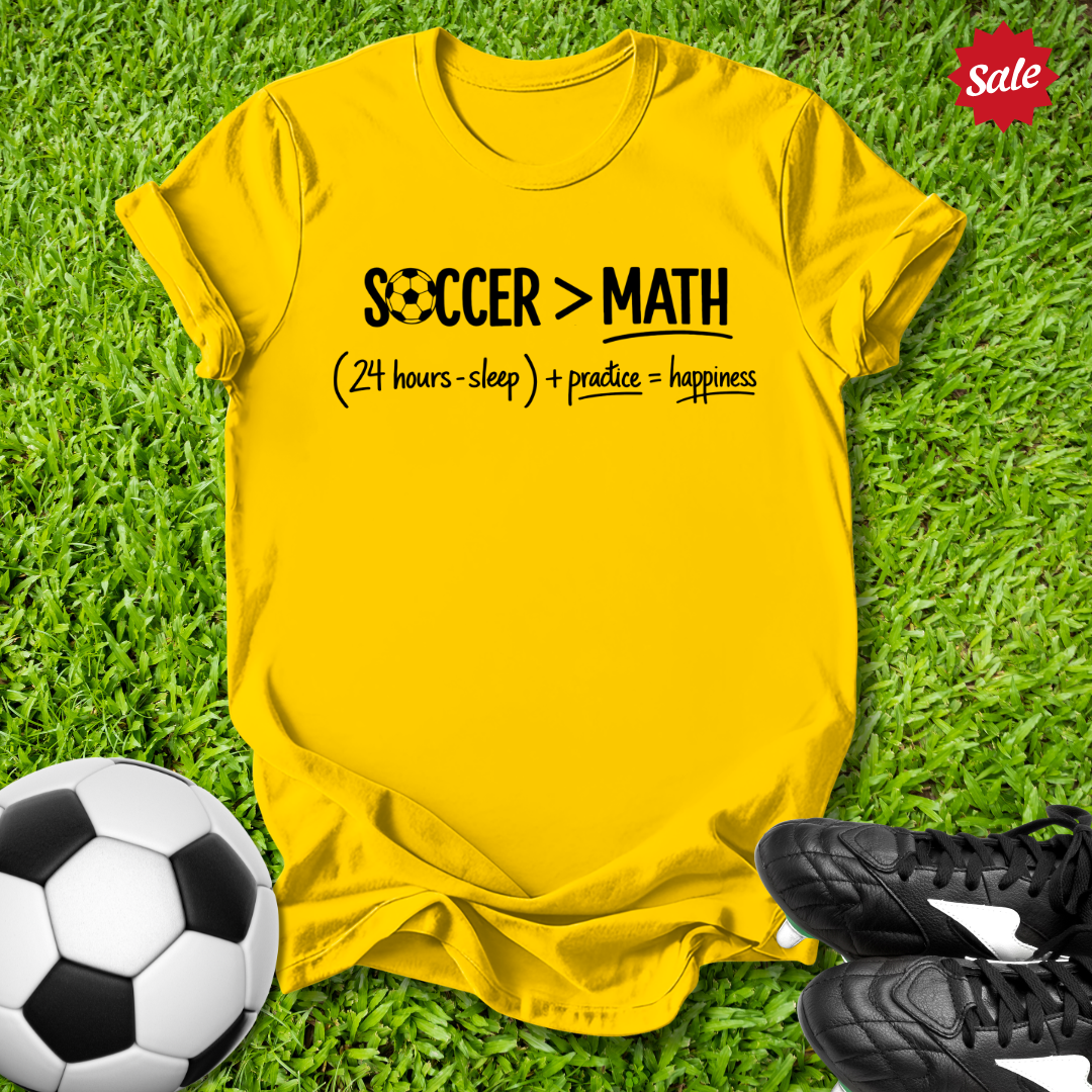 Yellow t-shirt with 'Soccer > Math' text on grass with soccer ball and shoes