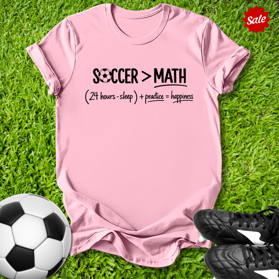 Pink t-shirt with 'Soccer > Math' text on grass with soccer ball and shoes