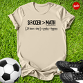 Beige t-shirt with 'Soccer > Math' text on grass with soccer ball and shoes